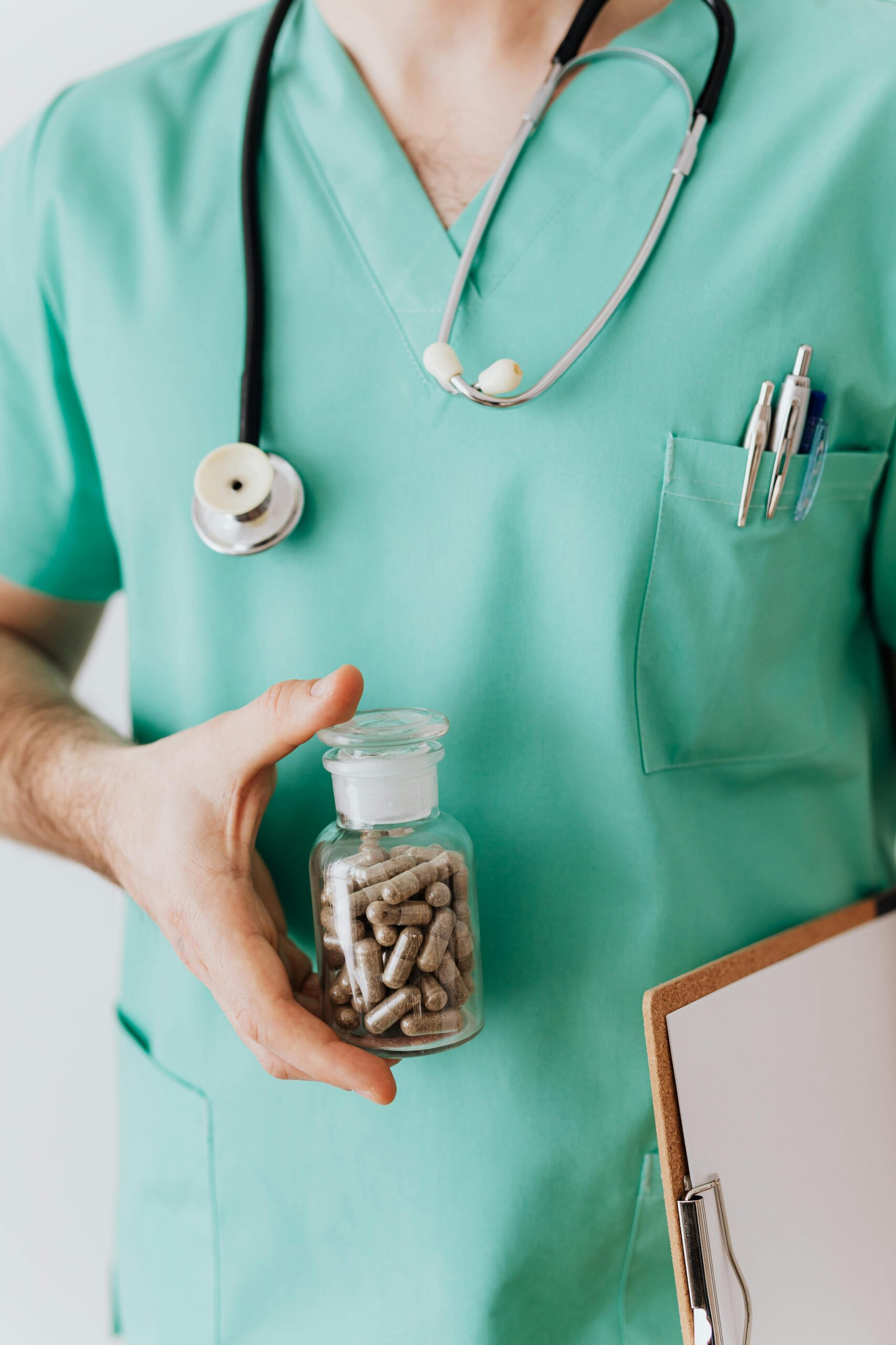 Faceless crop male doctor in medical uniform holding clipboard and demonstrating bottle with drugs while standing with stethoscope and pens in pocket